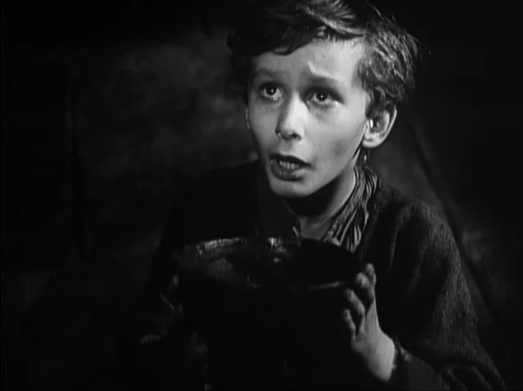 Oliver Twist (1948) movie still: closeup — A dimly lit, dark space with minimal architectural detail visible in the shadows. A young boy holds a…