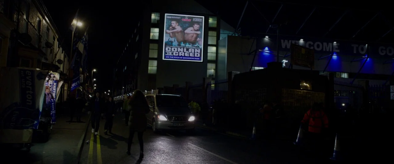 Creed (2015) movie still: wide — A dark city street at night runs alongside the exterior of Goodison Park stadium. A bright digital poster for a…