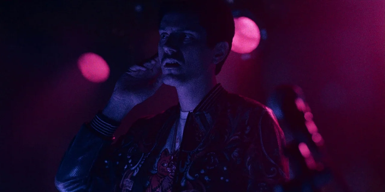 They Cloned Tyrone (2023) movie still: medium — A dark, atmospheric concert venue is bathed in deep magenta and purple neon light. A man wearing a…