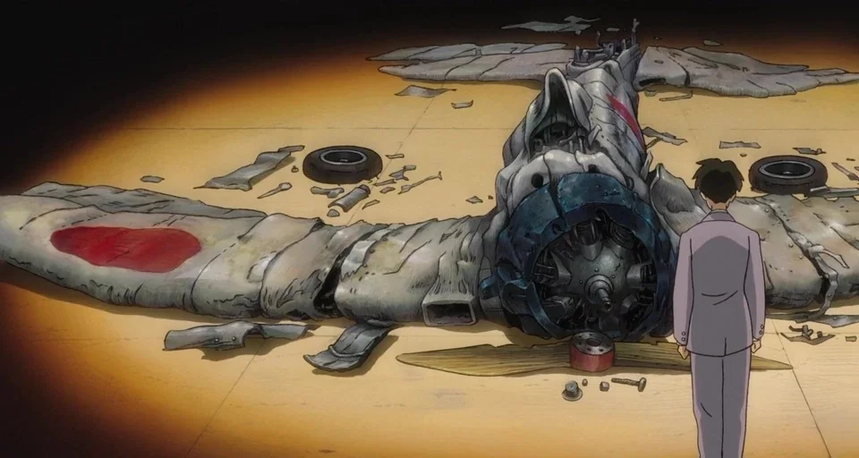 The Wind Rises (2013) movie still: wide — An expanse of wooden floorboards holds the mangled remains of a Japanese fighter plane. A man stands…