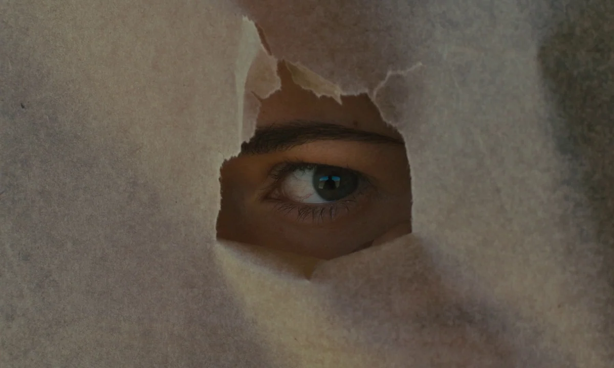 Slow West (2015) movie still: closeup — A dark, textured surface with a jagged irregular tear reveals an eye. The person behind the paper material…