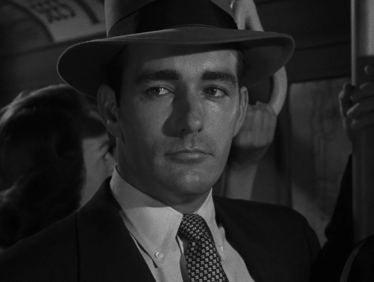 Pickup on South Street (1953) movie still: closeup — A man in a fedora rides on a public transit vehicle at night. He grips the metal handle of the…