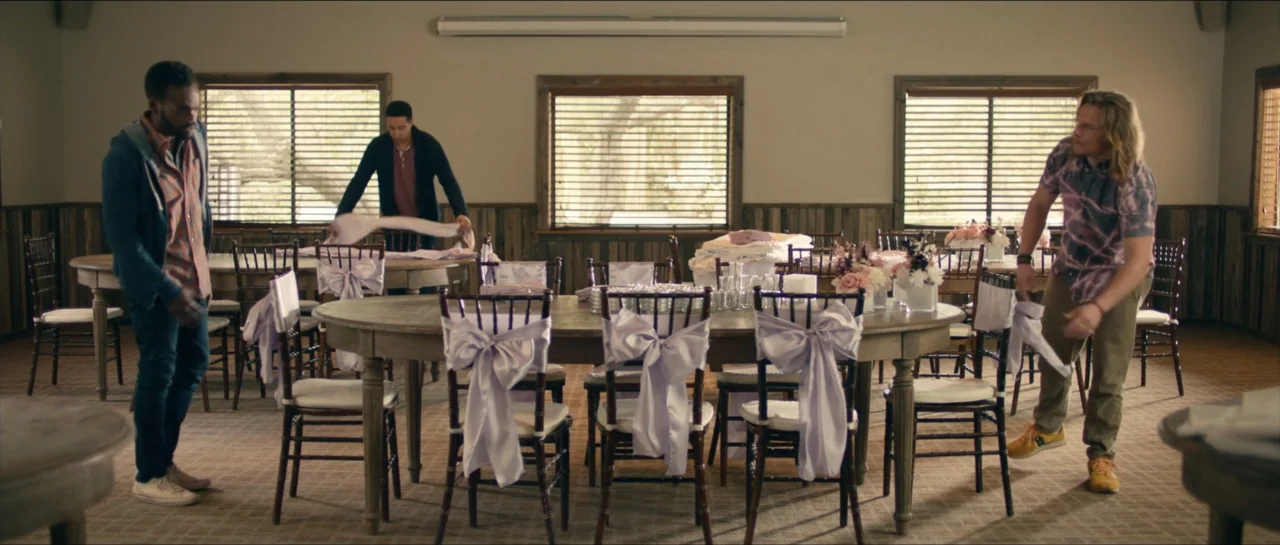 We Broke Up (2021) movie still: wide — A neutrally colored event hall contains several wooden tables adorned with purple sash-tied chairs. Three men…