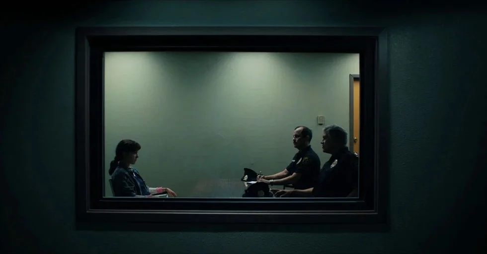 Stranger Things (2016) movie still: wide — A dark, dimly lit room with a large rectangular observation window at its center. Two police officers sit…