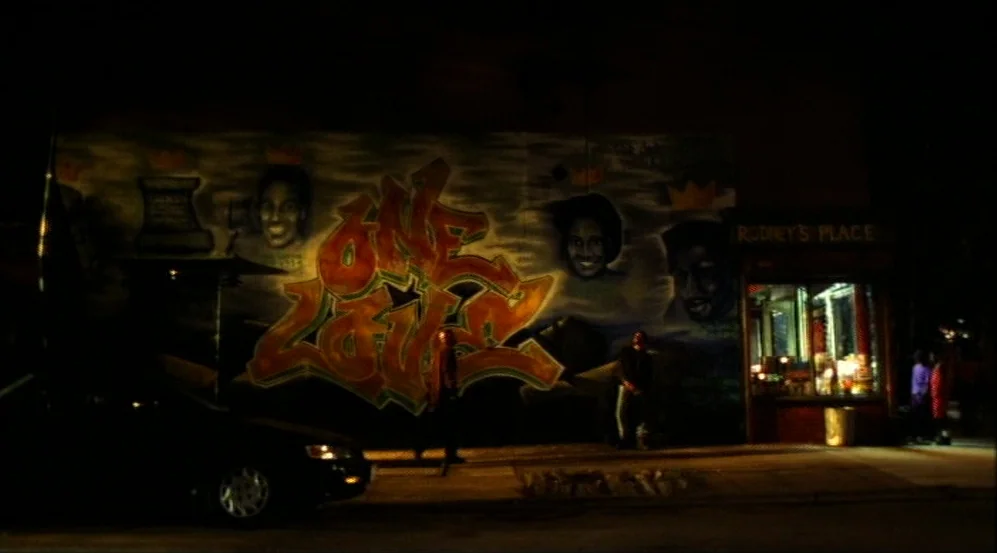 Clockers (1995) movie still: wide — A dark night exterior features a large wall adorned with stylized graffiti and portrait paintings. A small…