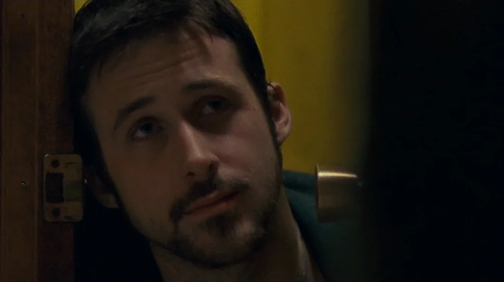 Half Nelson (2006) movie still: closeup — A man stands near a doorway with a yellow wall in the background. He leans against the wooden frame and…
