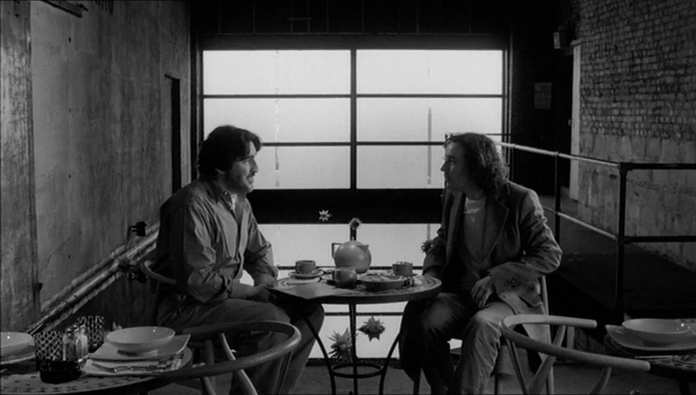 Coffee and Cigarettes (2003) movie still: medium — Two men converse at a small circular table situated against a large rear window. The room…