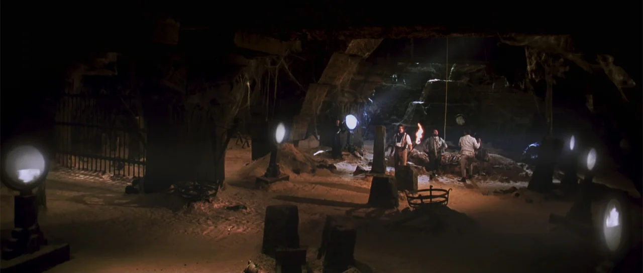 The Mummy (1999) movie still: wide — An ancient, sand-filled underground chamber features stone pillars and hanging cobwebs in deep shadow. Three…