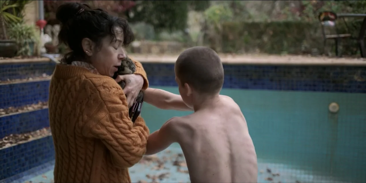 Bring Her Back (2025) movie still: medium — A woman and a shirtless boy stand inside a large empty tiled swimming pool. The woman holds the boy…