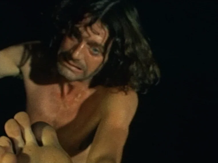 Salome (1972) movie still: closeup — A man without a shirt appears in a dark, undefined environment. He leans forward while extending his bare foot…