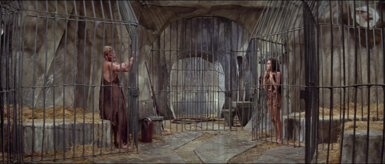 Planet of the Apes (1968) (1968) movie still: wide — A subterranean room features stone walls and several iron cages filled with straw. A man stands…