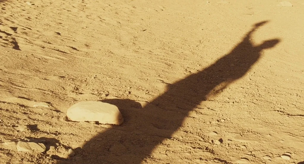 127 Hours (2010) movie still: wide — An arid, sun-baked landscape features sandy ground with scattered rocks and loose sediment. A long, dark shadow…
