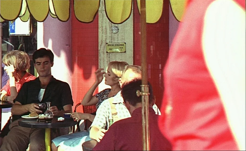 Le Bonheur (Happiness) (1965) movie still: medium — A sunlit street-side cafe terrace features a red-tiled wall with a yellow scalloped awning.…
