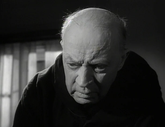 The Devil’s Eye (1960) movie still: closeup — A dark, dimly lit room contains an elderly man in a dark hood. The man looks downward while…