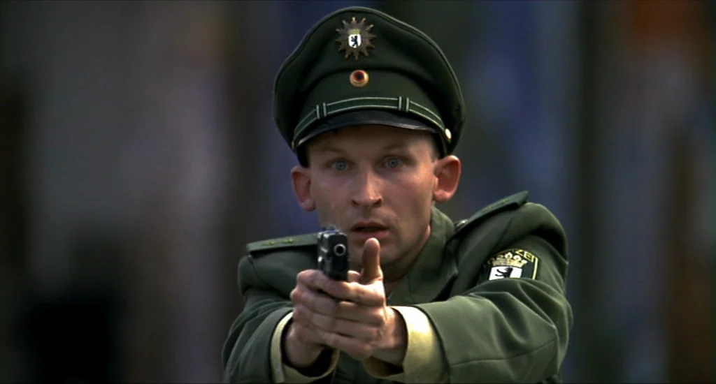Run Lola Run (1998) movie still: medium — A man in a German police uniform holds a handgun steady in both hands. He stands on an out-of-focus city…