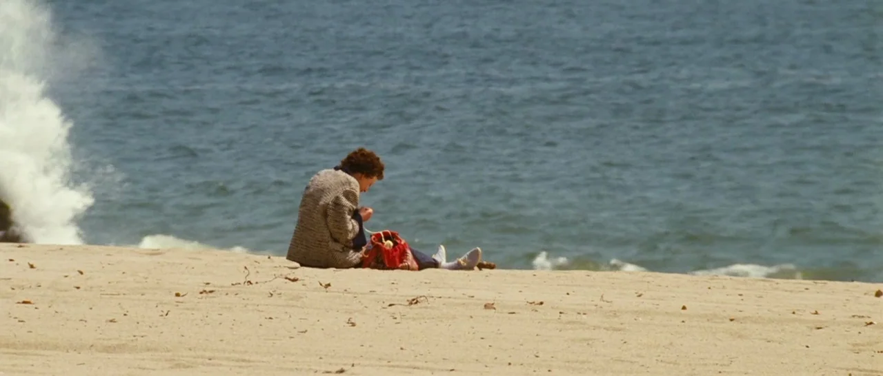 La Vie En Rose (2007) movie still: wide — A wide sandy beach features an ocean with breaking waves on the left. A woman sits on the sand facing the…