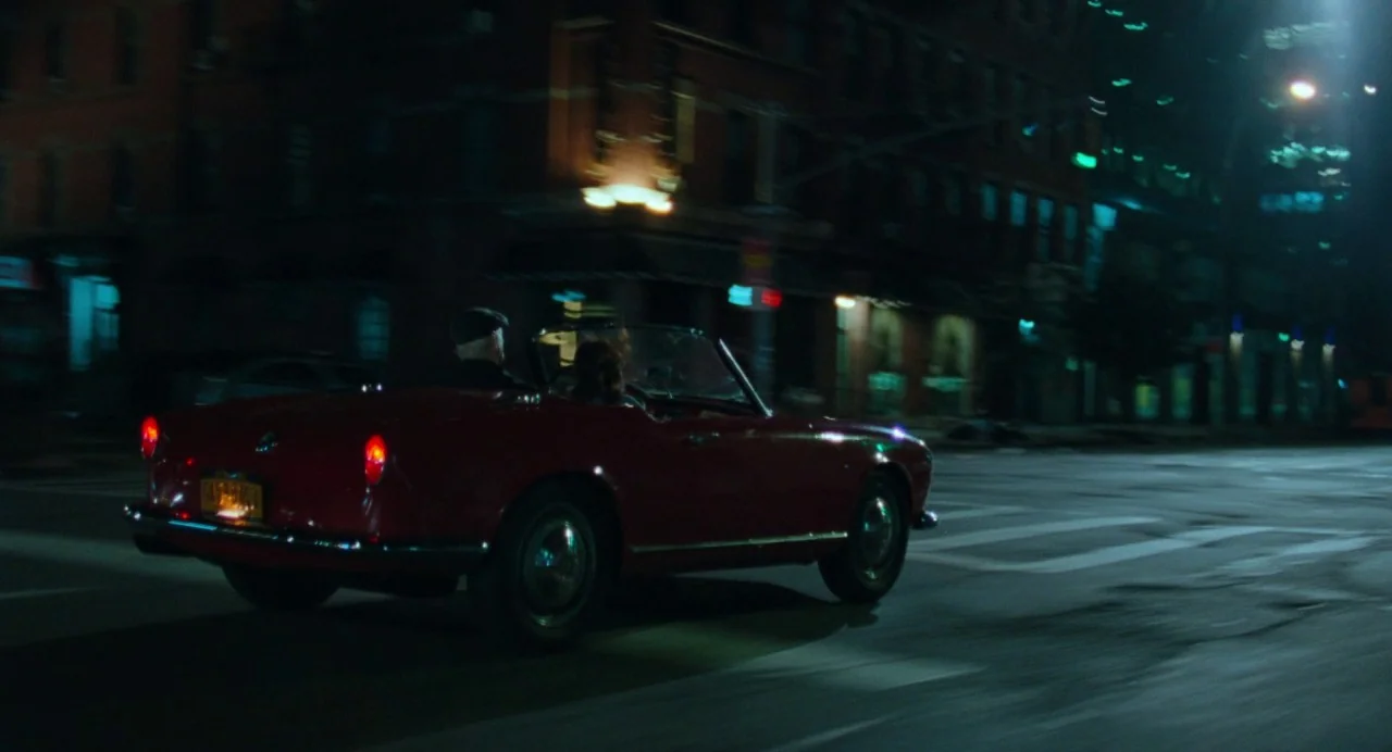 On The Rocks (2020) movie still: wide — A red convertible travels down a dark city street at night under dim lighting. Two people occupy the vehicle…