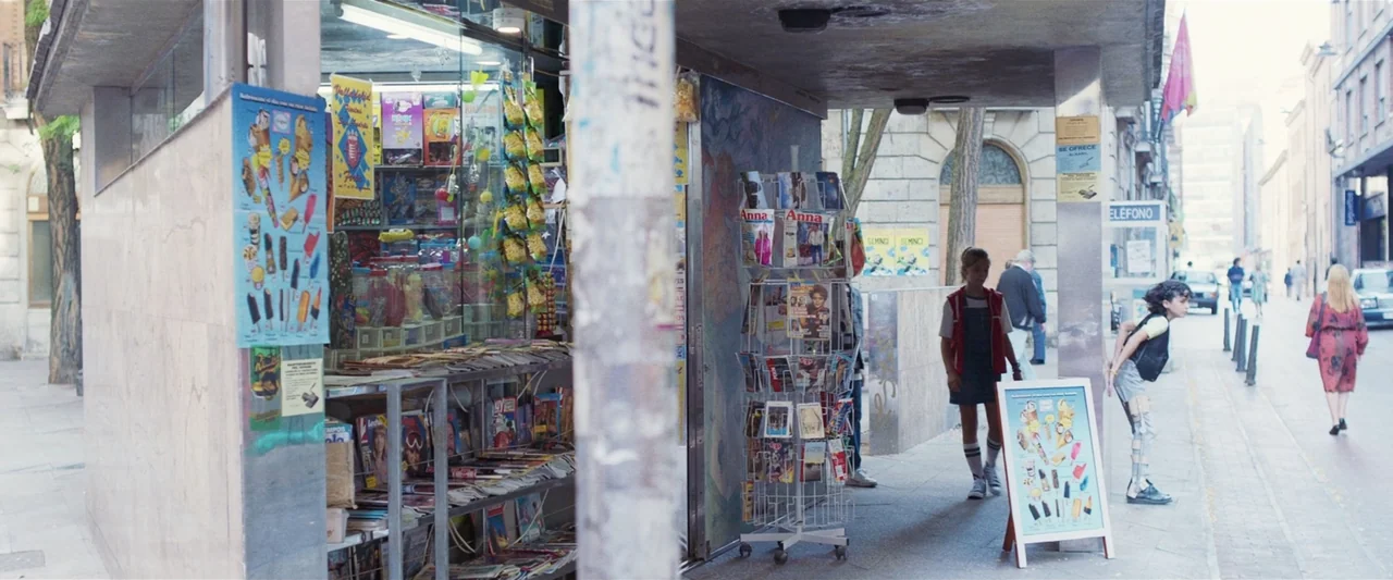 Voy a pasármelo bien (2022) movie still: wide — A city kiosk displays magazines and ice cream advertisements under a concrete overhang. A young girl…