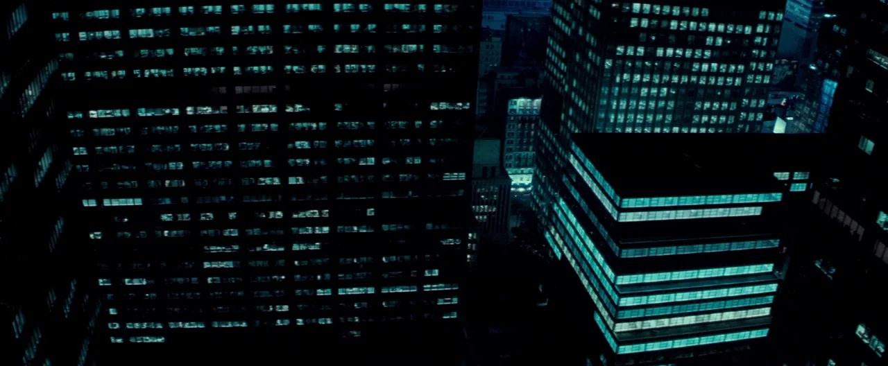 Michael Clayton (2007) movie still: establishing — A dense urban nightscape features dark skyscrapers with rows of illuminated teal office windows.…