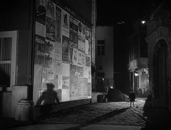 The Third Man (1949) movie still: wide — A narrow cobblestone street at night is lined with buildings covered in advertisement posters. A small lone…