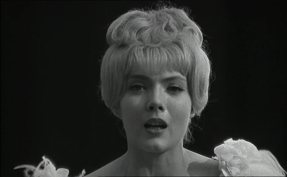 Cléo from 5 to 7 movie still: closeup — A dark stage with no visible background elements surrounds a single female singer. The woman is centered,…