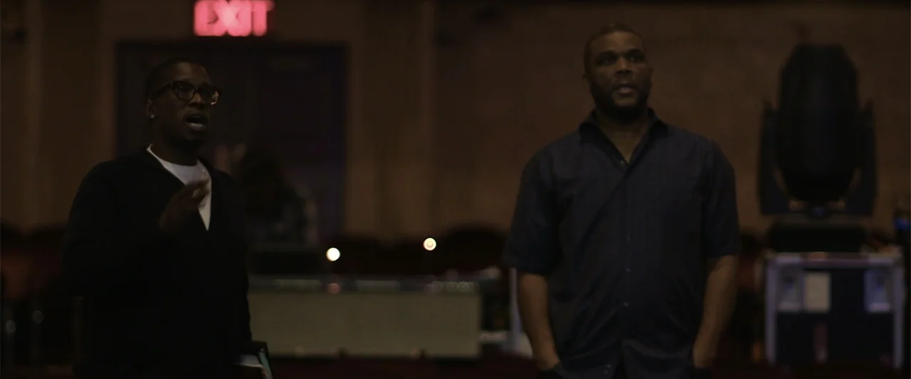 Maxine's Baby: The Tyler Perry Story (2023) movie still: medium — A dark empty auditorium with rows of seating and a glowing red exit sign. Two men…
