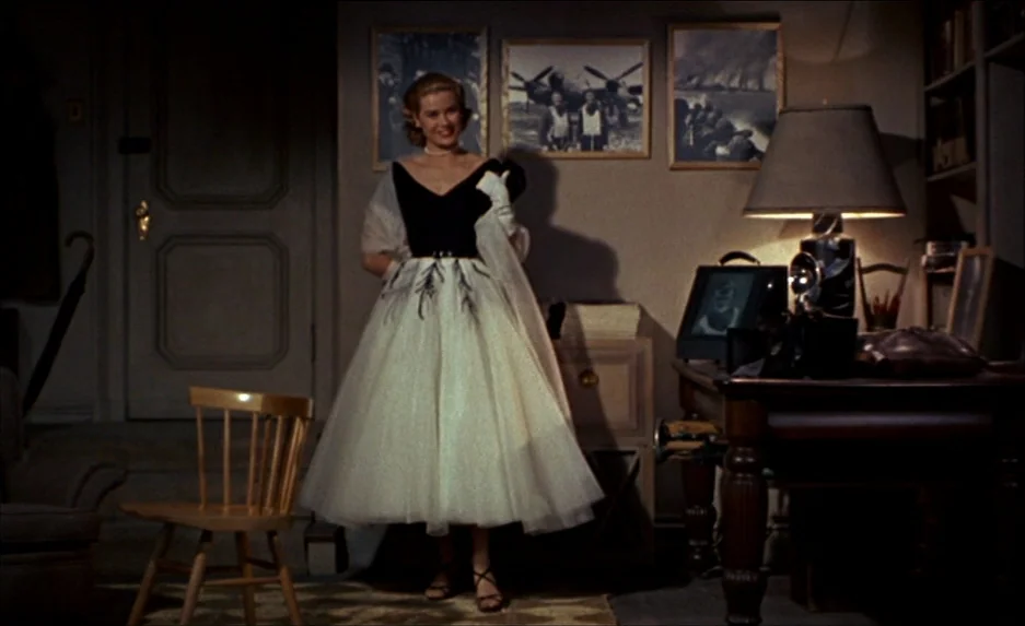 Rear Window (1954) movie still: fullbody — A dimly lit home study contains a dark wood desk, framed photographs, and a chair. A woman in a black and…