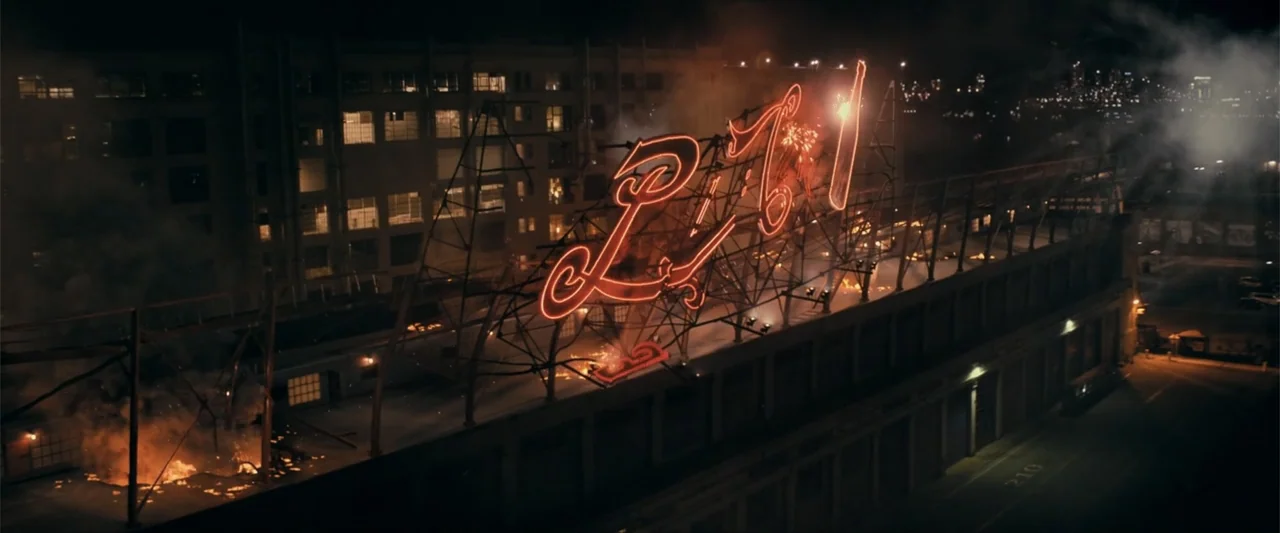 Madame Web (2024) movie still: wide — A multi-story industrial building features a massive red neon sign on its roof. Intense fire erupts on the…