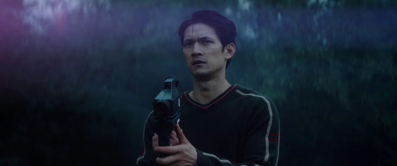 Broadcast Signal Intrusion (2021) movie still: medium — A man stands in an outdoor area at night surrounded by dark blurred foliage. He holds a…