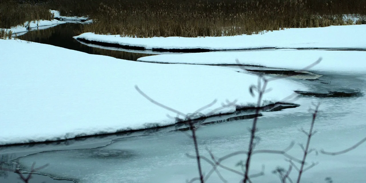 Polar (2019) movie still: establishing — A winding river is partially covered in thick white snow and jagged ice formations. Tall dry brown reeds…