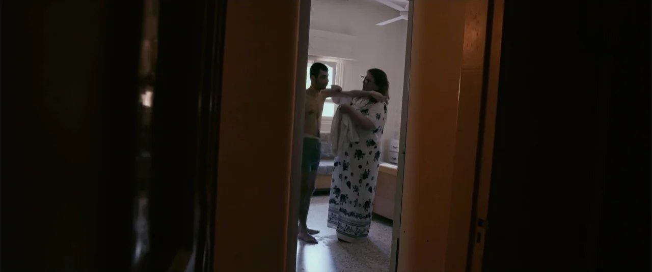 Simple as Water (2021) movie still: medium — A sparsely furnished room features a window behind a man and a woman. The woman stands opposite the man…