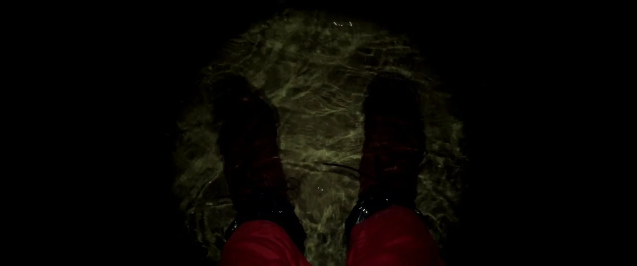 The House That Jack Built (2018) movie still: closeup — A dark, nocturnal setting reveals feet standing in shallow, rippling water. A beam of light…