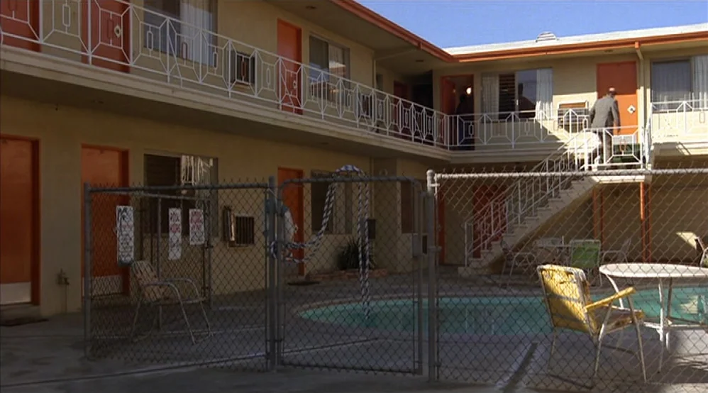 Hardcore (1979) movie still: establishing — A two-story stucco motel courtyard features orange doors and a fenced pool area. A man in a suit walks…