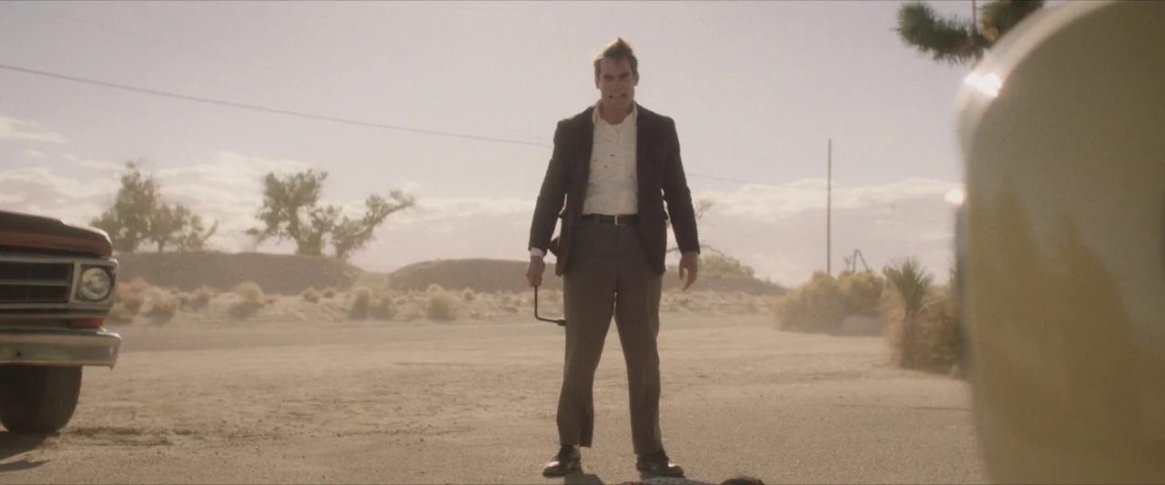 The Last Stop in Yuma County (2023) movie still: fullbody — A man stands in the middle of a dusty desert road between two vehicles. He holds a metal…