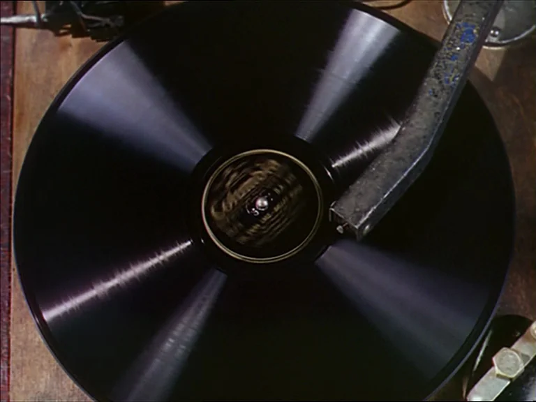 The Red Shoes (1948) movie still: closeup — A top-down view of a vintage record player turntable with a spinning black disc. The metal tonearm is…