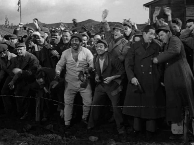 Stalag 17 (1953) movie still: wide — A group of prisoners stands behind a barbed wire fence in a dirt lot. The men cheer and wave their arms toward…