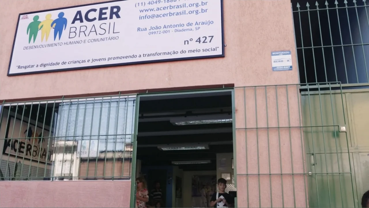 Finding the Way Home (1991) movie still: wide — A light-colored building exterior features a large rectangular sign for Acer Brasil above the door.…