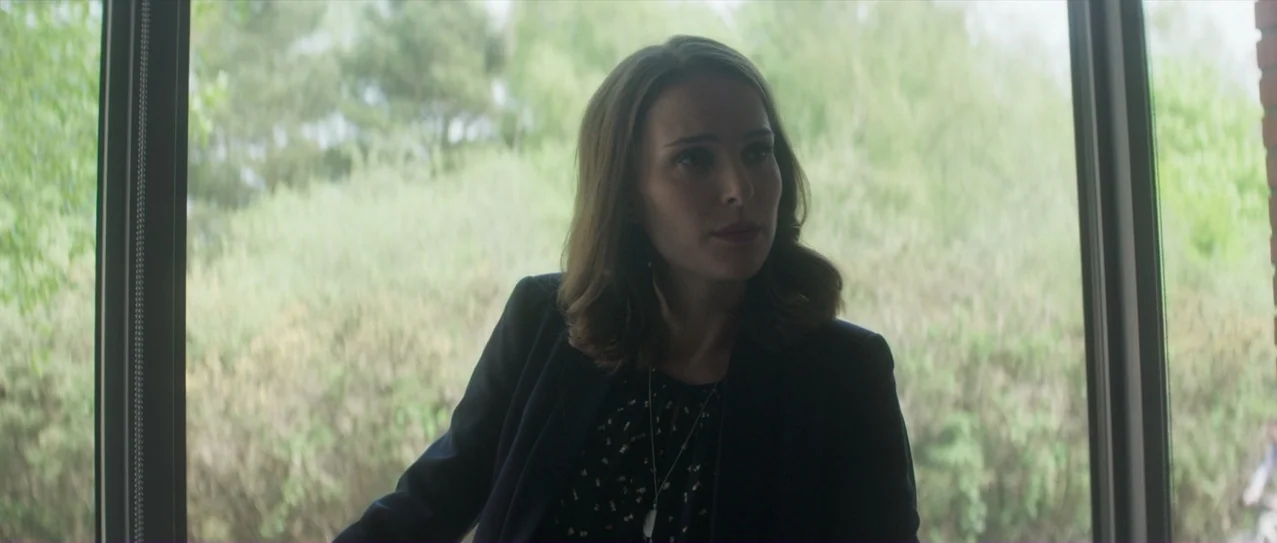 Annihilation (2018) movie still: medium — A woman stands indoors in front of large glass windows facing a green garden. The subject looks to the…