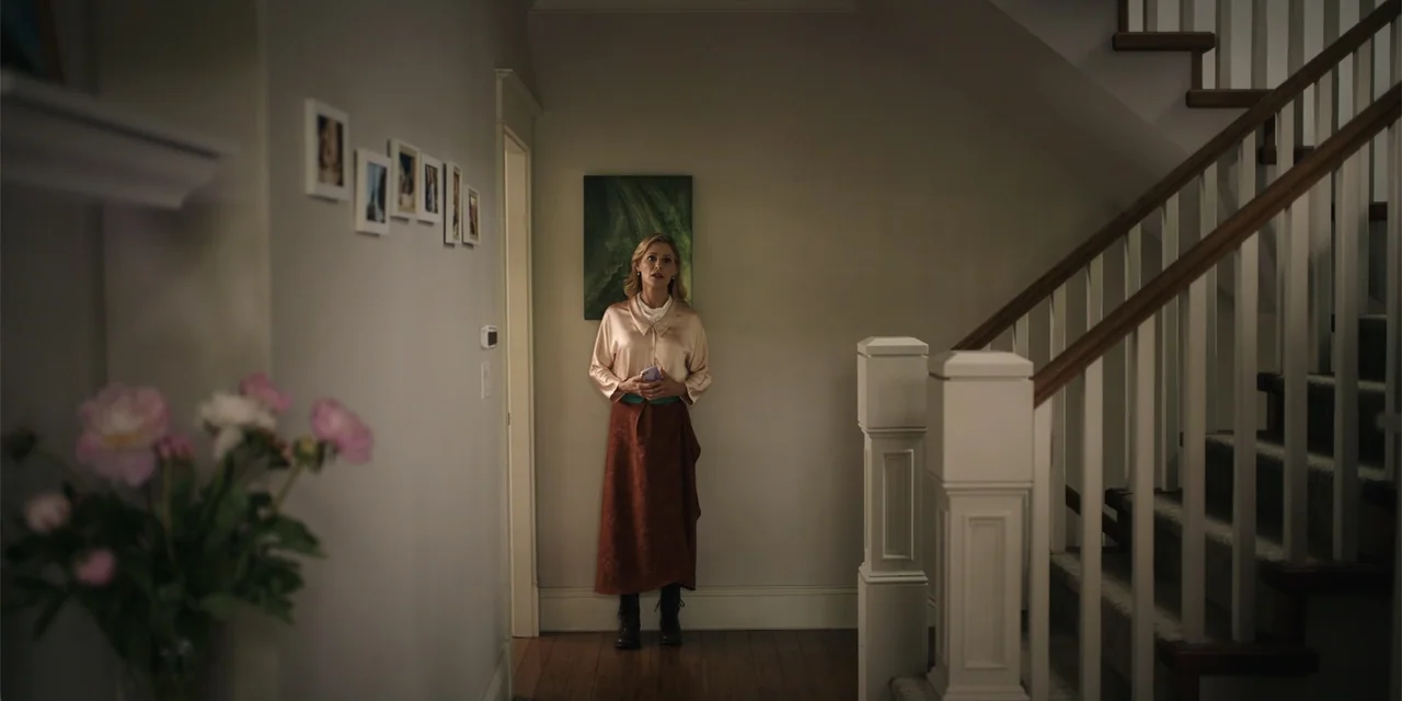 Totally Killer (2023) movie still: fullbody — A residential hallway with wooden floors, light walls, and a staircase to the right. A woman stands in…