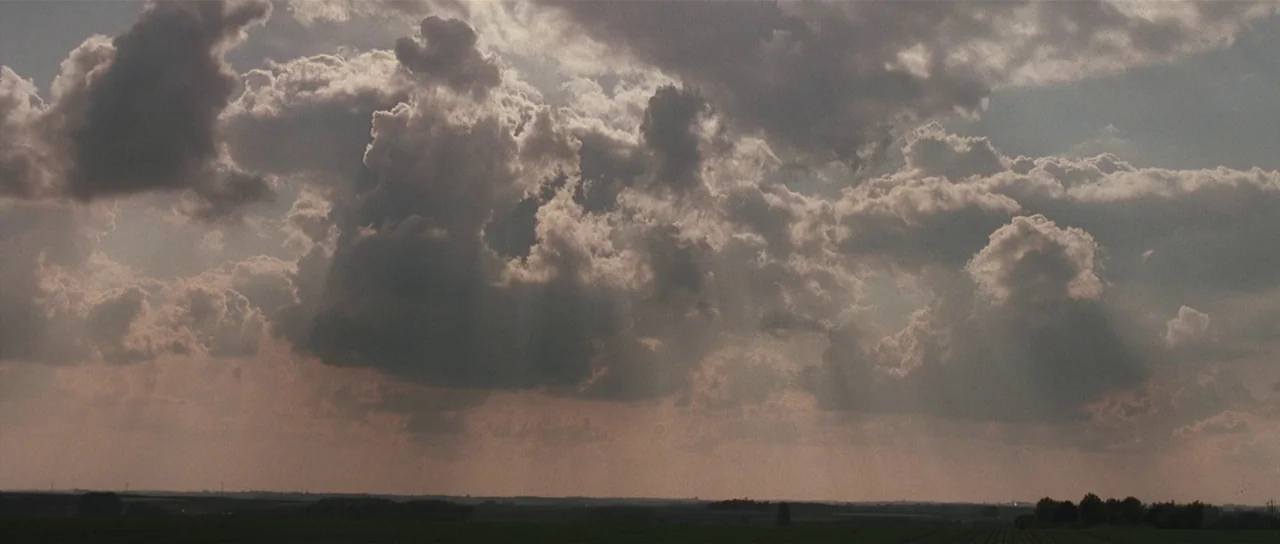 Bullhead (2011) movie still: establishing — A vast, flat rural field stretches under a dramatic, heavily clouded afternoon sky. Sunlight breaks…