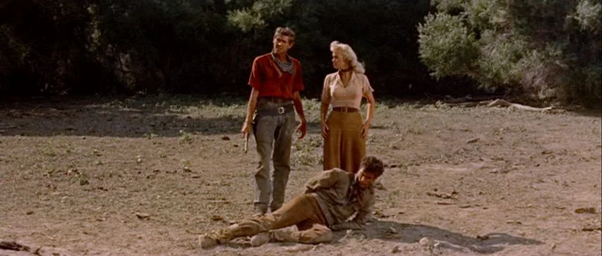 Ride Lonesome (1959) movie still: fullbody — A dry desert clearing surrounded by scrub brush and low trees. One man stands with a gun while a woman…