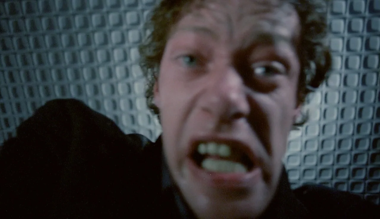 Angst (1983) movie still: closeup — A close-up shot of a man's face in front of a textured wall. The subject stares directly at the lens with an…