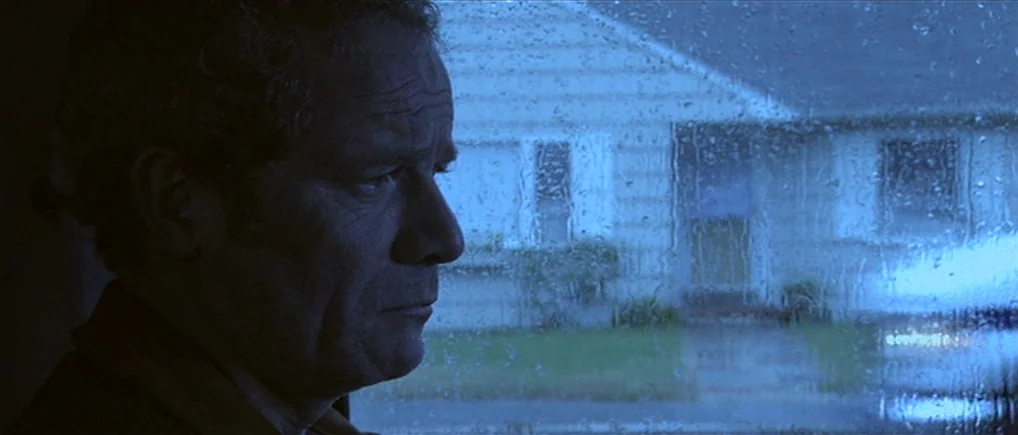 Session 9 (2001) movie still: closeup — An interior space faces a rain-covered window looking toward a suburban house exterior at night. A…