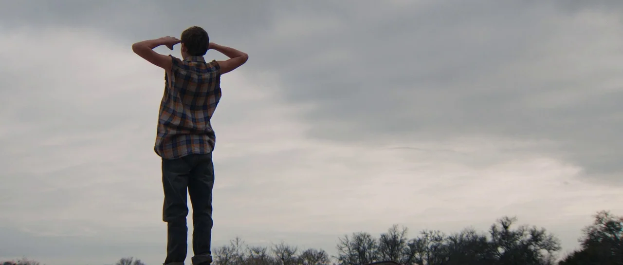 Joe (2013) movie still: fullbody — A figure stands in a vast, open landscape under a heavy overcast gray sky. The person faces away, shielding their…