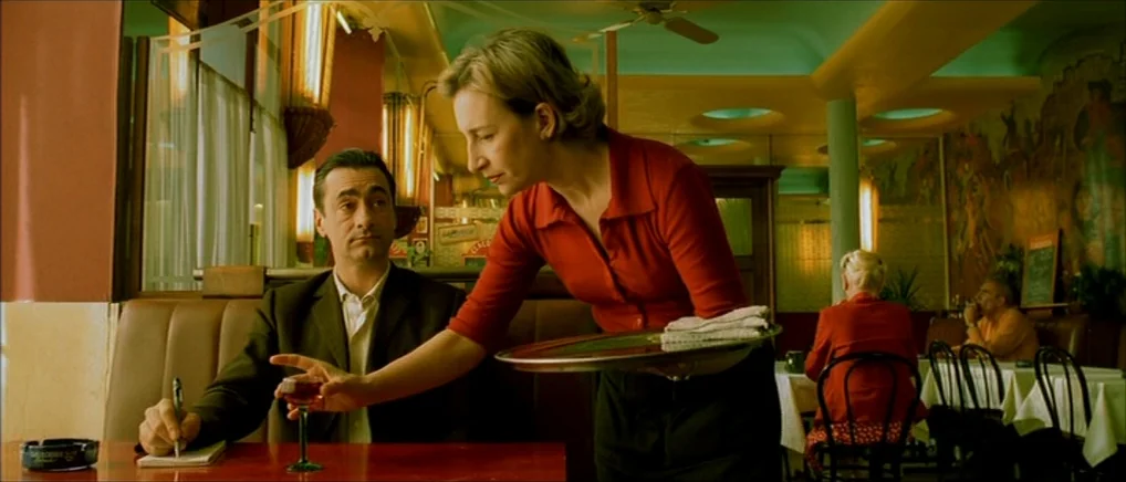Amelie (2001) movie still: medium — A diner interior features red booths and tables with a yellow-tinted wall mural. A blonde waitress in a red…