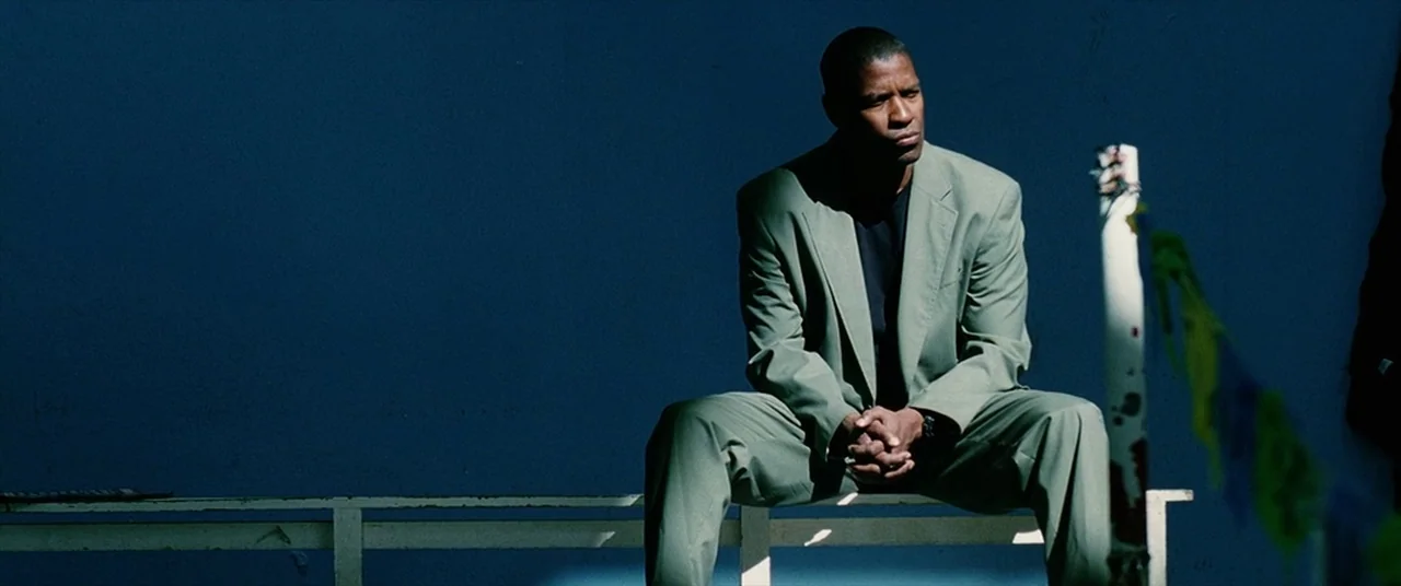 Man on Fire (2004) movie still: medium — A man sits on a white bench against a dark blue solid background wall. He is wearing a light green suit and…