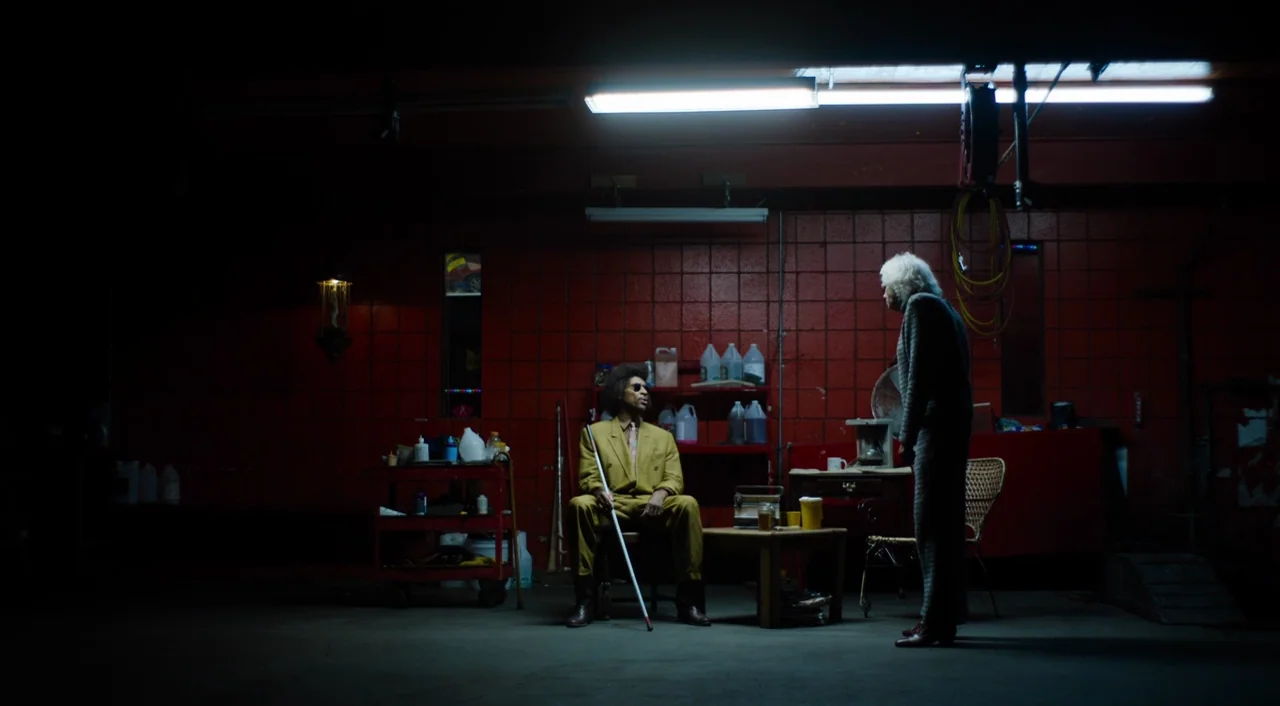 The Greasy Strangler (2016) movie still: wide — A dimly lit garage with red brick walls contains various chemical containers and tools. A man seated…