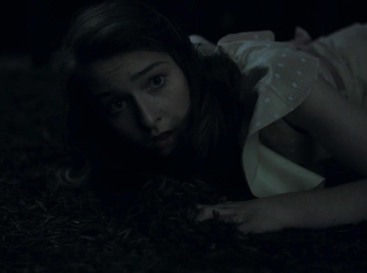 Girl Asleep (2015) movie still: closeup — The subject lies on dark, textured earth in a low-light outdoor setting. A young woman in a patterned…