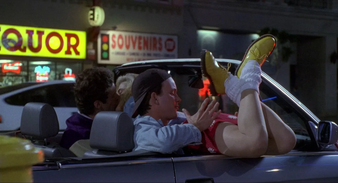 Buffy the Vampire Slayer (1992) movie still: medium — A city street at night features a convertible car parked in front of shops. A couple sits…