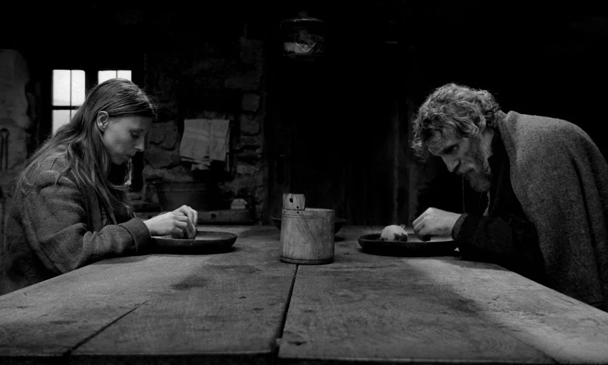 The Turin Horse (2011) movie still: medium — A dimly lit room with stone walls contains a long heavy wooden table. A man and a woman sit facing each…